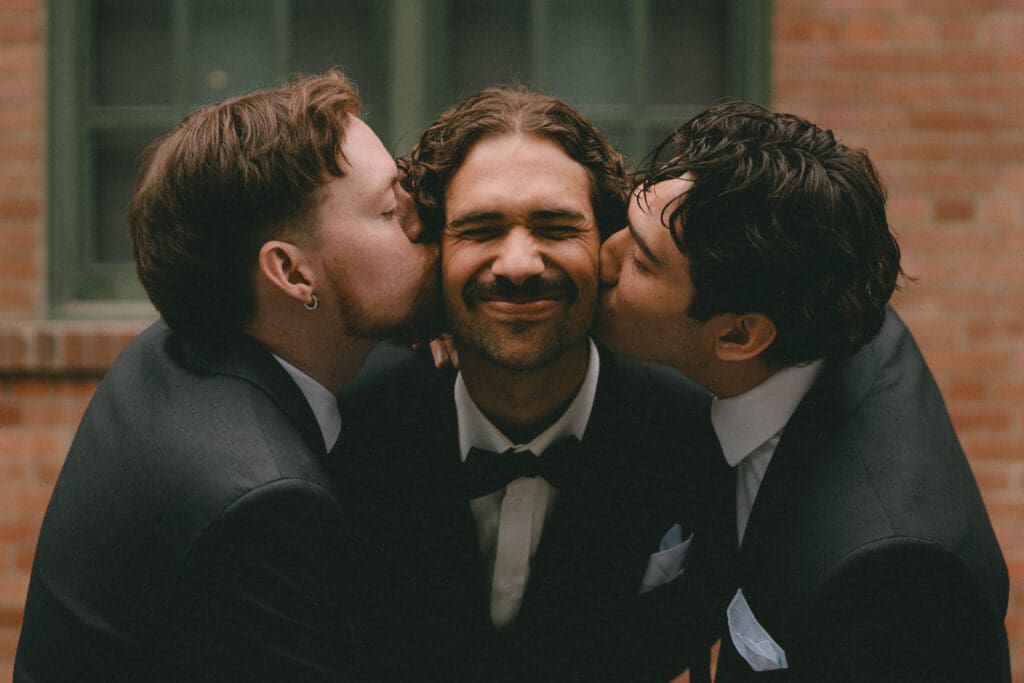 Groomsmen kissing groom on the cheek in front of brick building