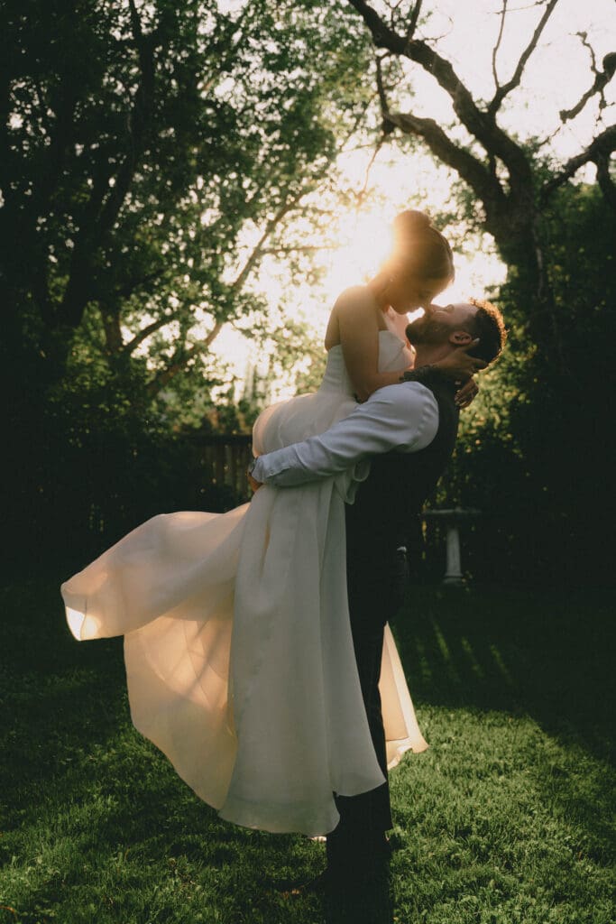 Groom picking up bride in front of some trees during sunset