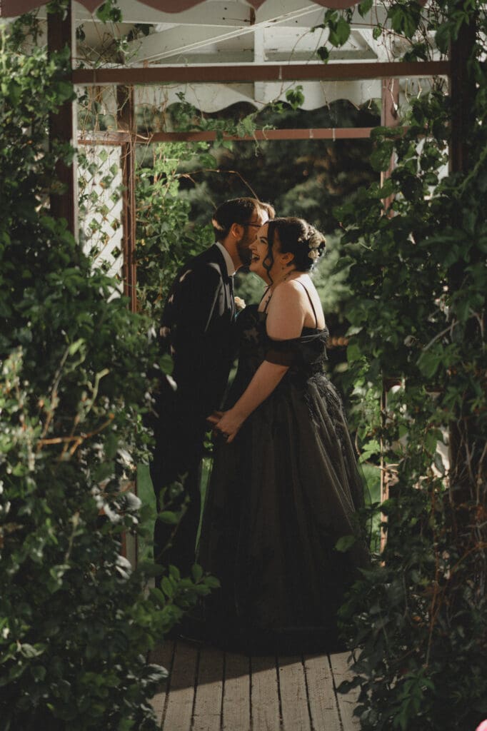 Bride in black dress laughing at groom whispering in her ear under a gazebo